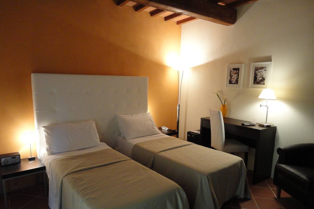 Una habitación de hotel con dos camas individuales, un escritorio, una silla, una mesita de noche y una lámpara.