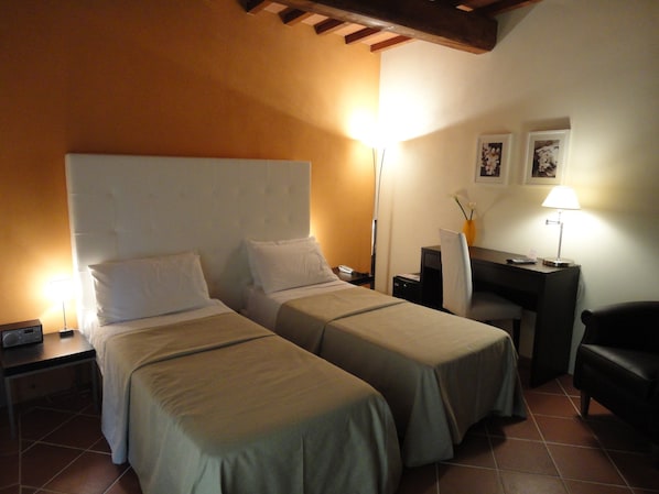 Habitació d'hotel amb dos llits individuals, una taula de treball, una cadira, una petita taula de nit i una làmpada.