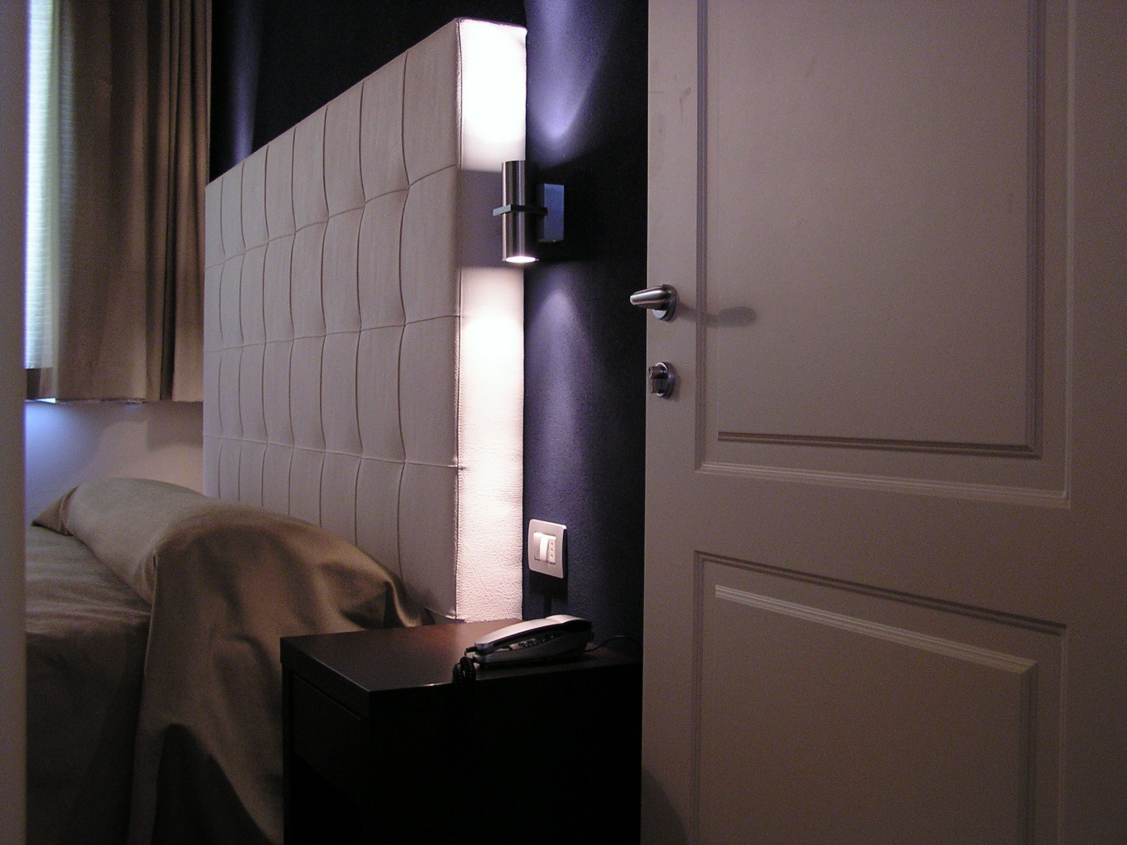 A hotel room with a bed, a bedside table with a telephone, and a door.