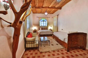 A bedroom with a bed, a wooden headboard, a nightstand, a chair, a small table, and a large window with curtains. - Riad Dar Etto (Essaouira)