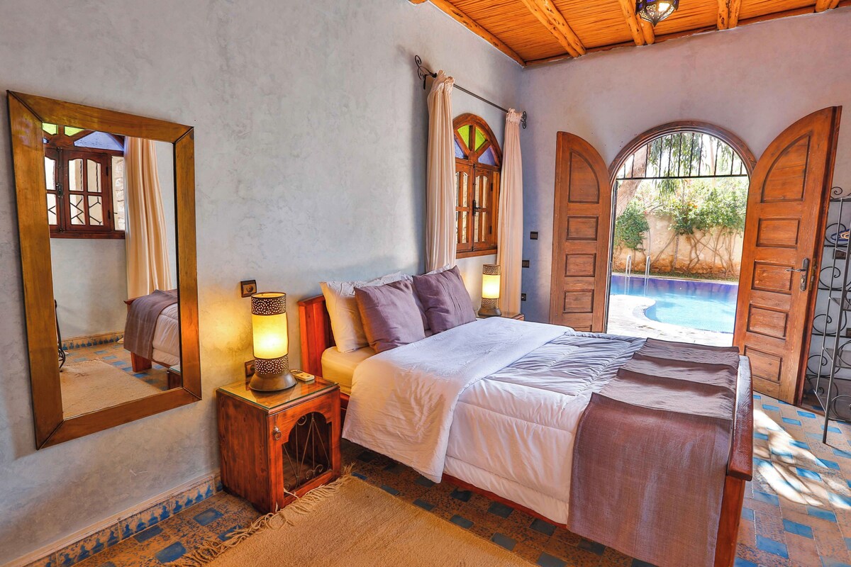 Un dormitorio con cama, mesita de noche, lámpara y un espejo grande. Hay una ventana con cortinas y vista a una piscina al aire libre.