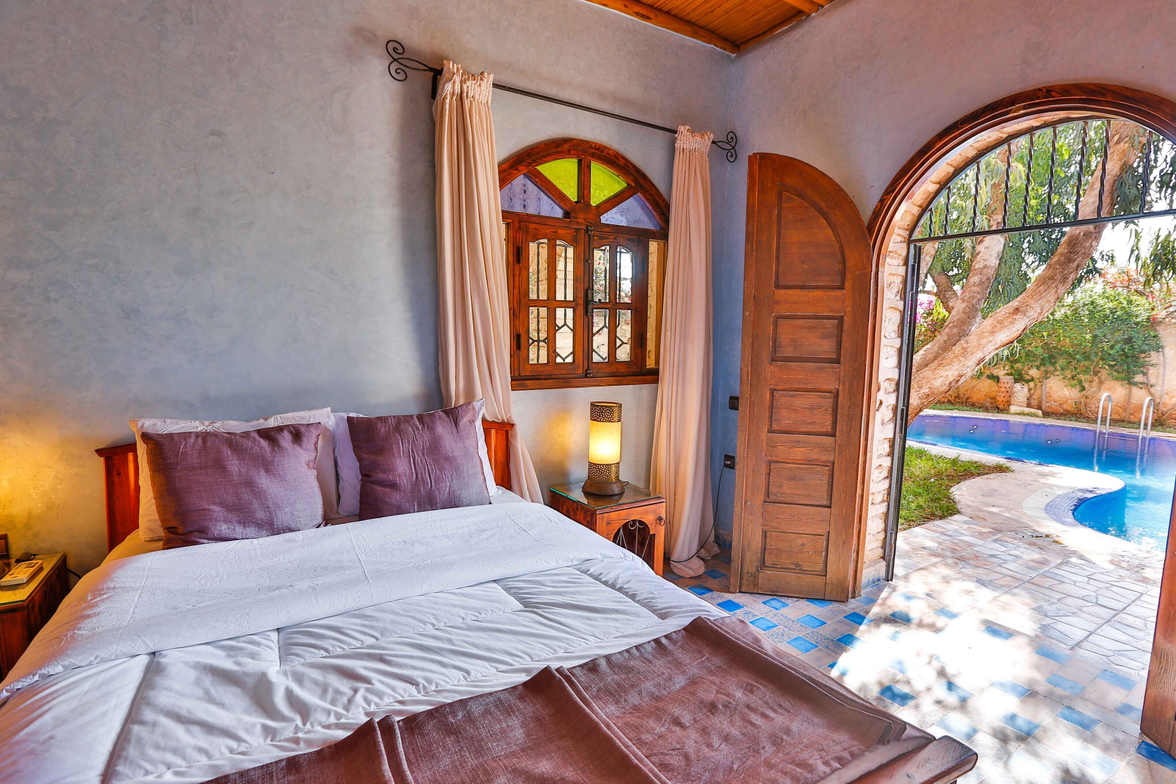 A bedroom with a bed, a nightstand, a lamp, a window with curtains, and a door leading to a pool area.
