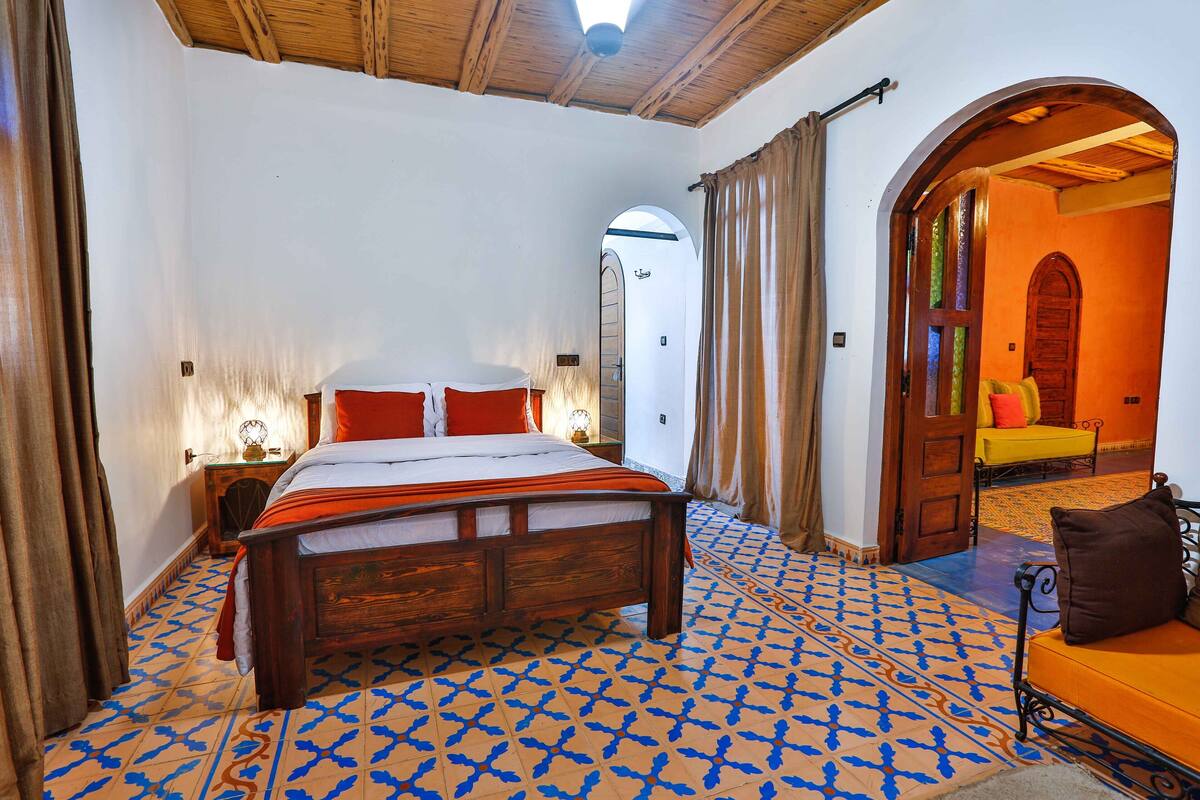 A bedroom with a wooden bed, patterned floor, and a view of a dining area through an arched doorway.