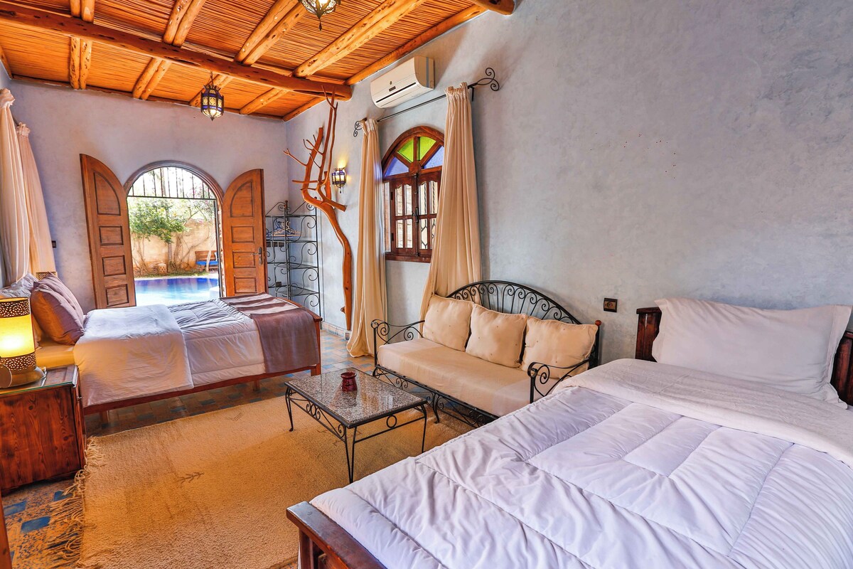 Una habitación con dos camas, techo de madera, una mesita y vista a una piscina.