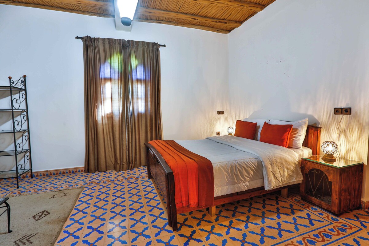 Un dormitorio con cama, mesita de noche, lámpara, estante y ventana con cortinas.