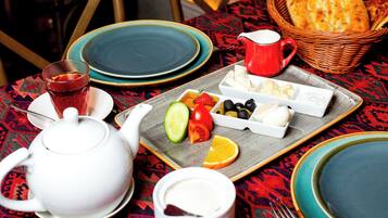 Una mesa dispuesta con un calentador de té, una taza, un plato con pan, queso y fruta, y una jarra de leche.