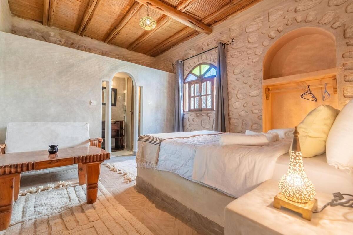 A cozy bedroom with a stone wall, a wooden bed, a nightstand with a lamp, and a small table with a chair.