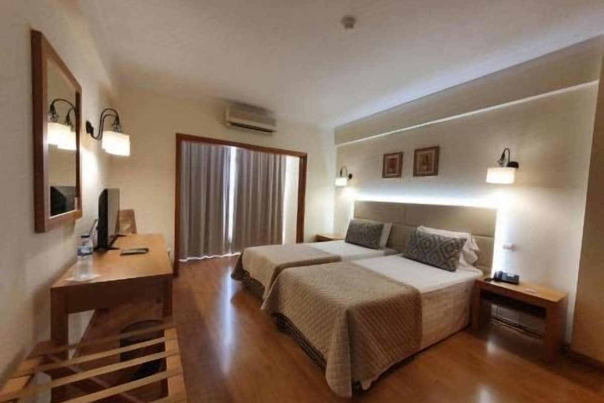 Quarto de hotel com duas camas, uma mesa, uma cadeira e uma janela com cortinas.