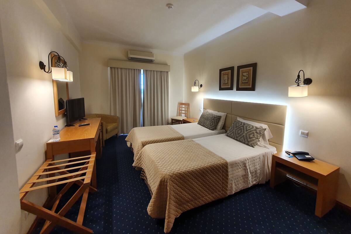 A hotel room with two beds, a desk, a chair, a small table, and a wardrobe.