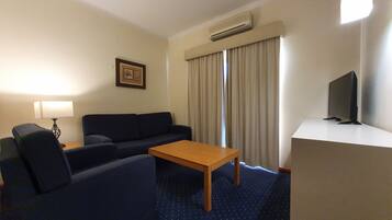 A hotel room with a sofa, a small table, a TV on a stand, a lamp, and a framed picture on the wall.
