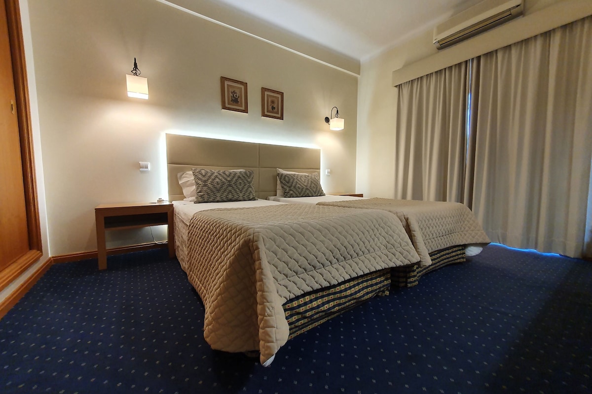 A hotel room with a large bed, a nightstand, a wall-mounted lamp, and a window with curtains.