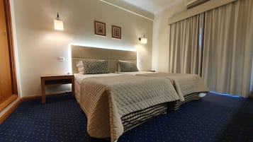 A hotel room with a large bed, a bedside table, a nightstand, a wall-mounted lamp, and a window with curtains.