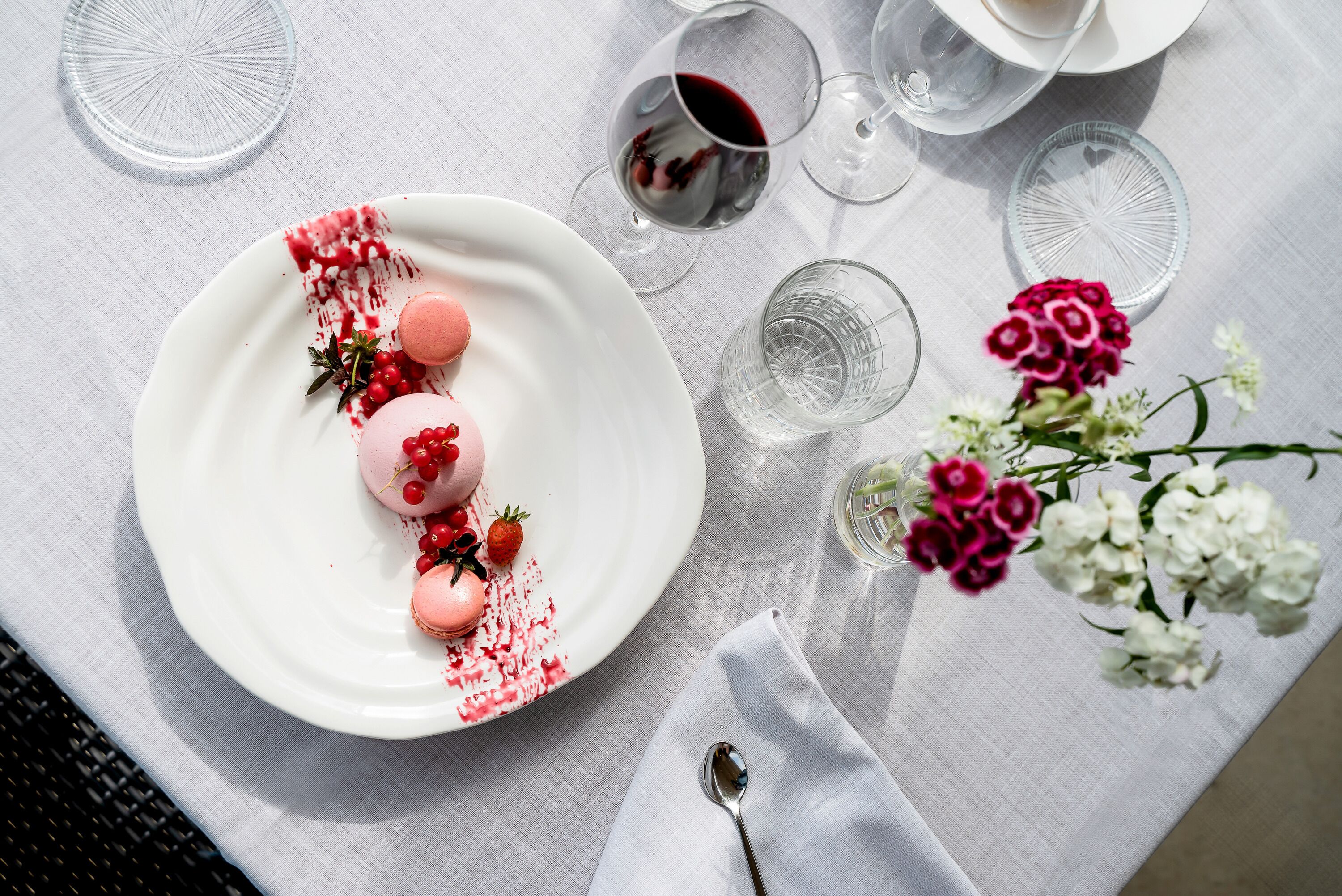 Una mesa con un plato blanco de macarons, una copa de vino tinto y un jarrón de flores.