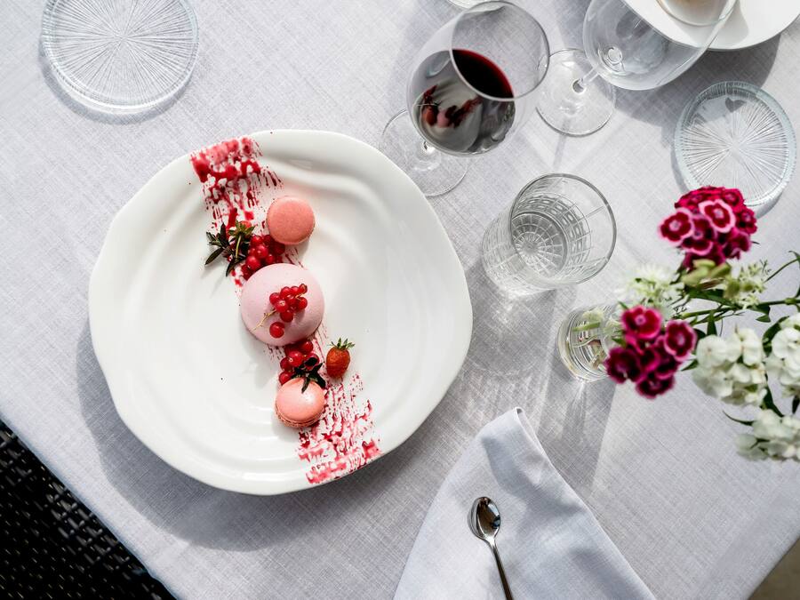 Uma mesa posta com um prato branco de macarons, um copo de vinho tinto e um vaso de flores.