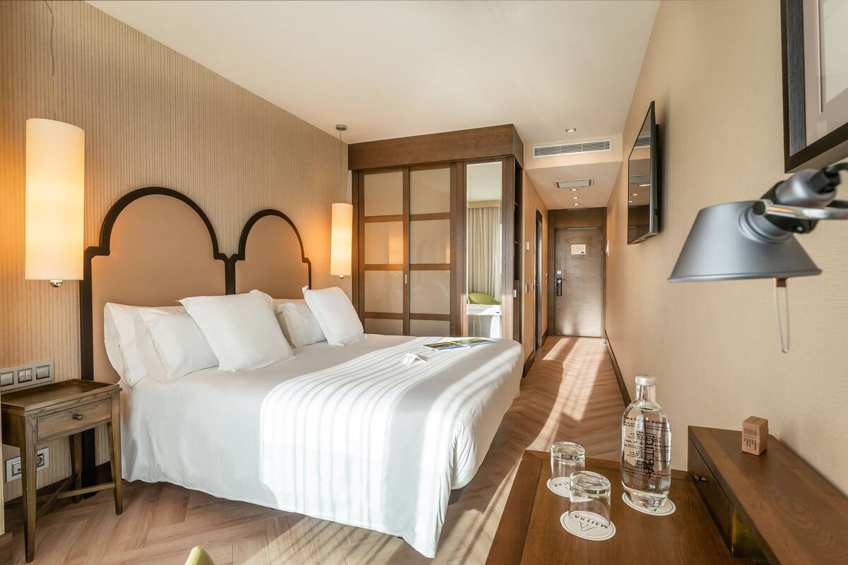 A hotel room with a large bed, bedside lamps, a wooden nightstand, a bedside table with a bottle and glasses, and a mirror on the wall.