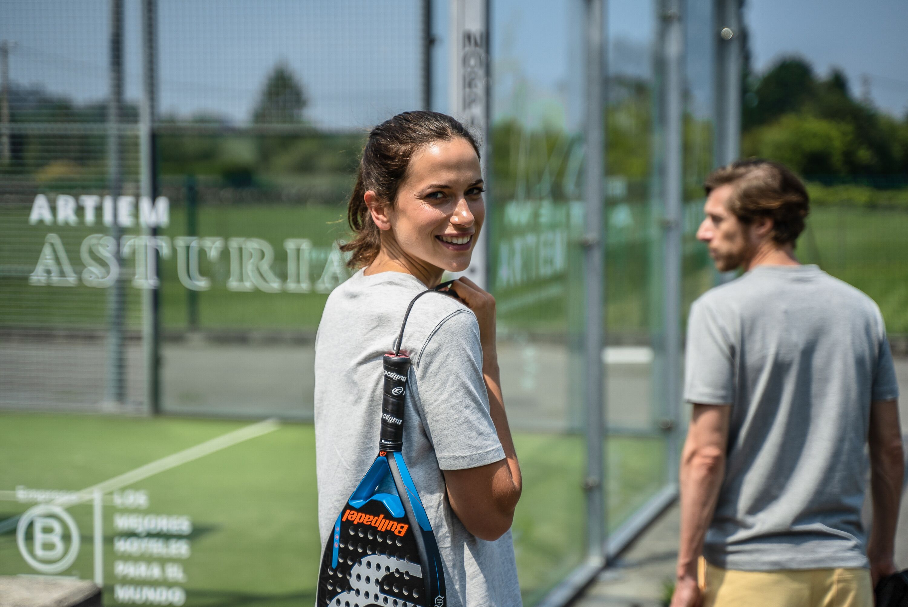 Henkilö seisoo urheiluhallissa, ja hänellä on mukanaan tennismelalaukku.