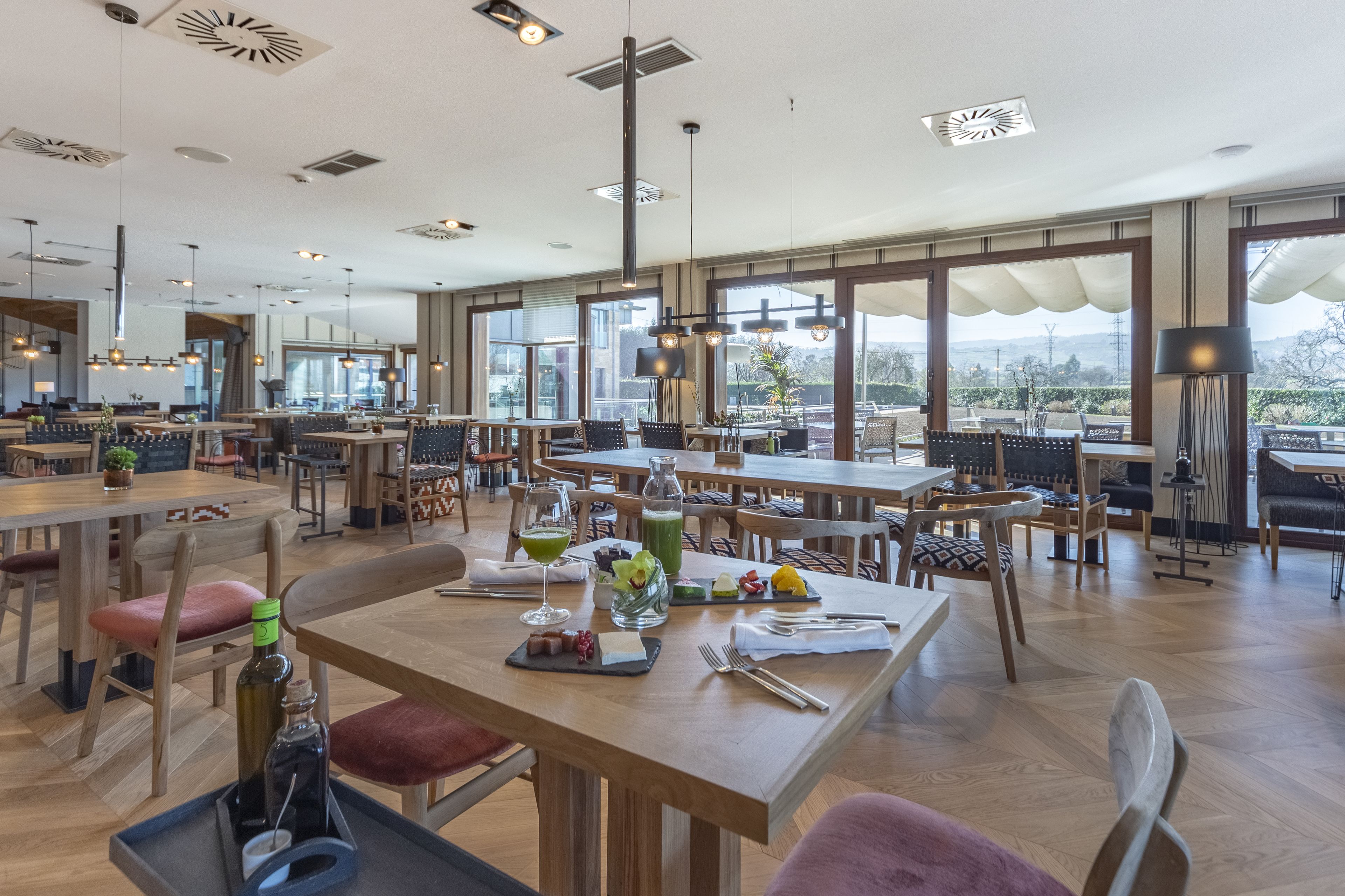 Un comedor amplio con varias mesas y sillas, ventanas grandes que ofrecen vistas al exterior, e iluminación colgante moderna.