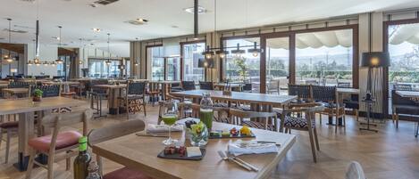Un comedor amplio con varias mesas y sillas, ventanas grandes que ofrecen vistas al exterior, e iluminación colgante moderna.