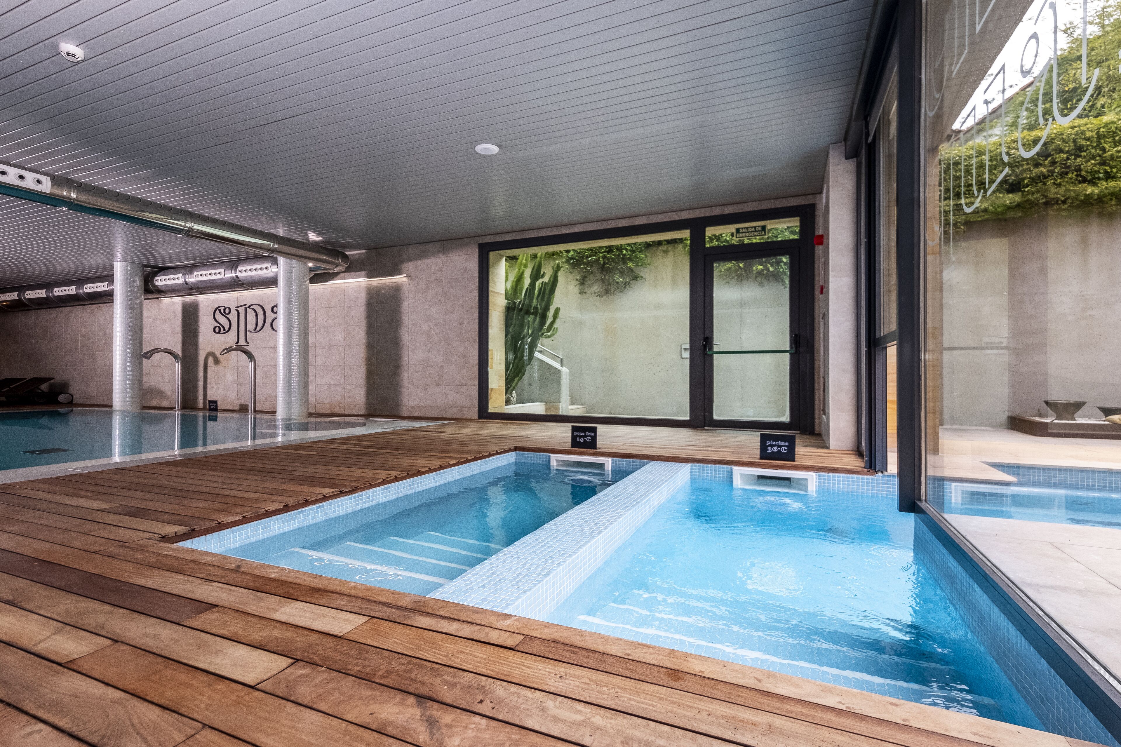 An indoor swimming pool with wooden decking and clear blue water, surrounded by a modern interior with a glass wall and a door leading outside.