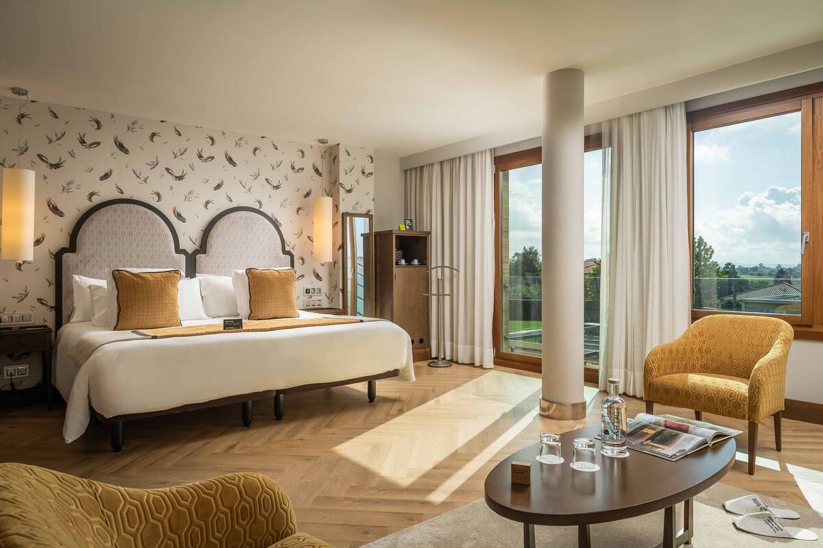 A hotel room with a large bed, a sitting area with a table, a chair, and a view of the outdoors.