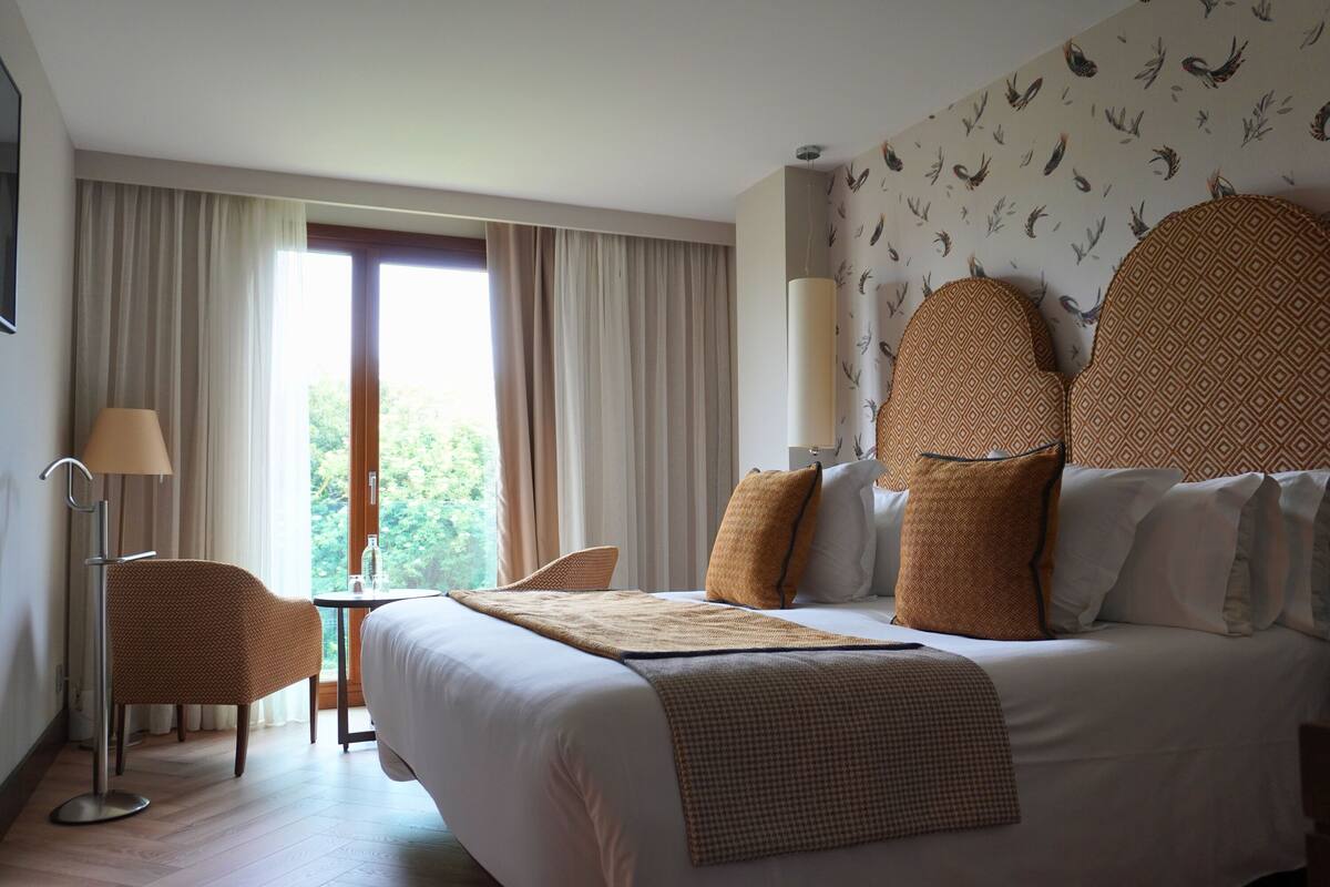 A hotel room with a large bed, a desk, a chair, and a view of greenery through the window.