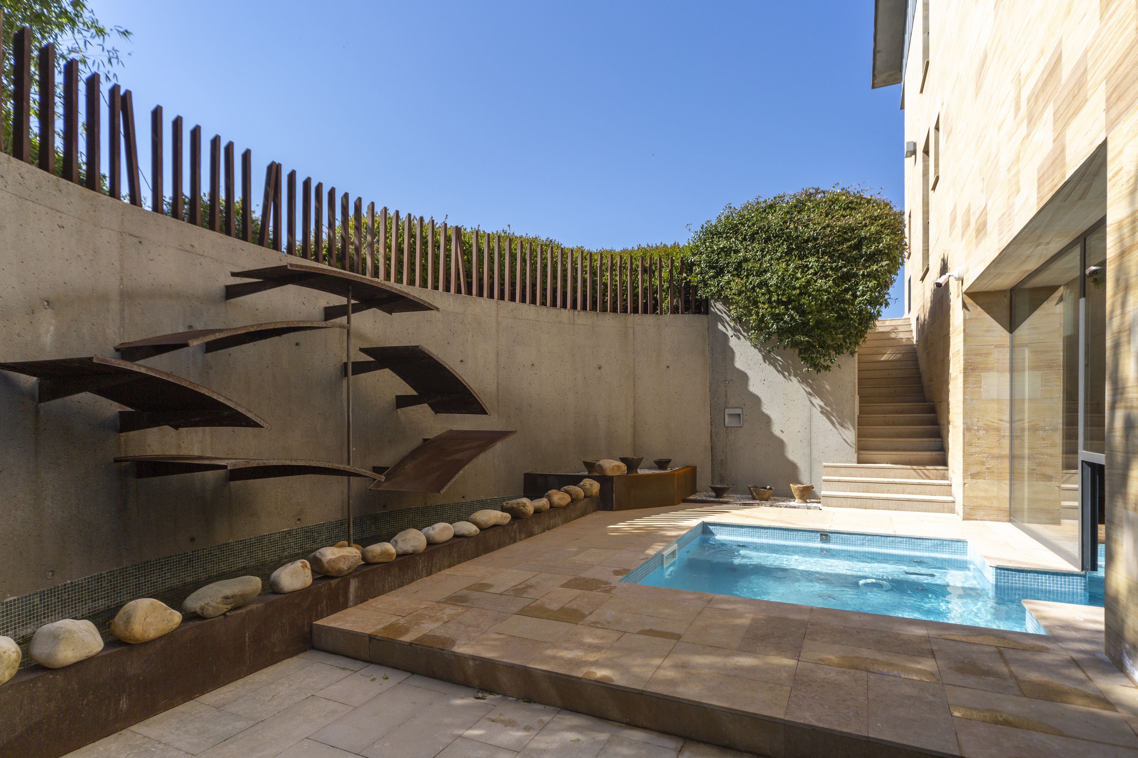 Un área de piscina moderna al aire libre, con un muro de hormigón, una valla de madera y una escalera que lleva a un edificio.