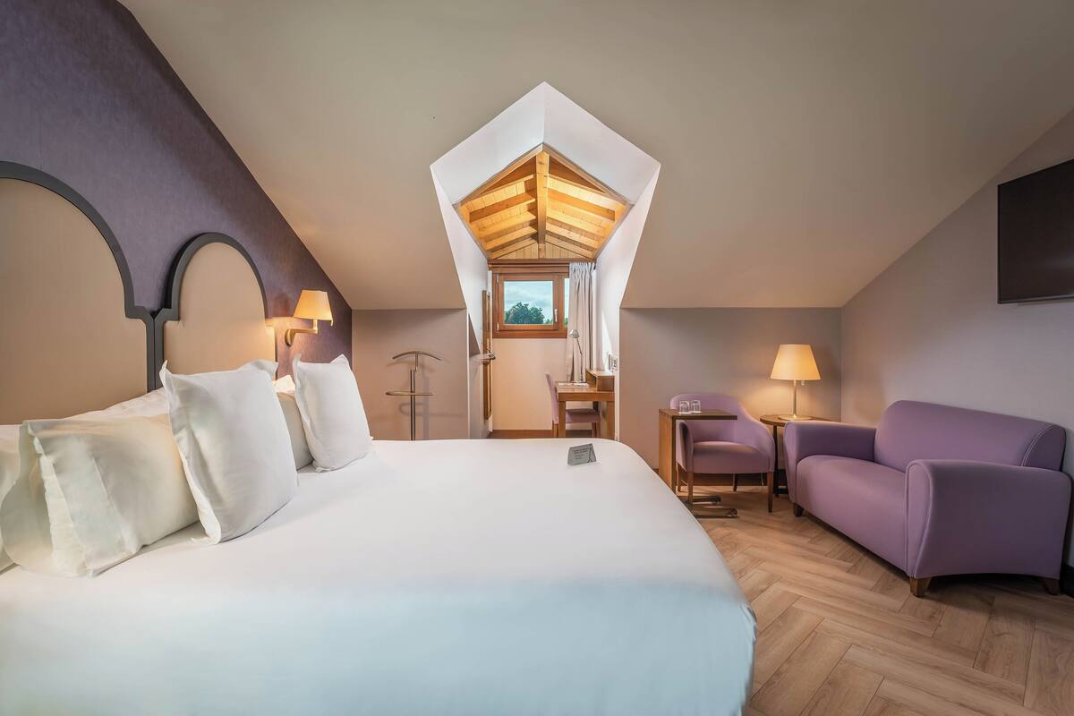 A bedroom with a large bed, a purple armchair, a desk, and a window with a view.