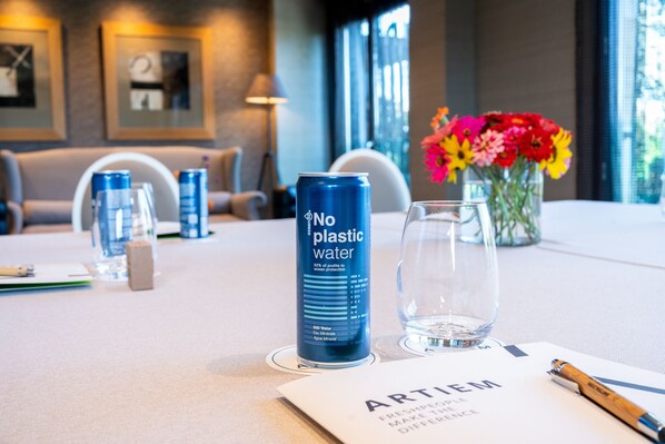 A hotel room with a table set for a meeting, featuring a can of 'No Plastic Water', a glass of water, a vase with flowers, and a document titled 'ARTIUM'.