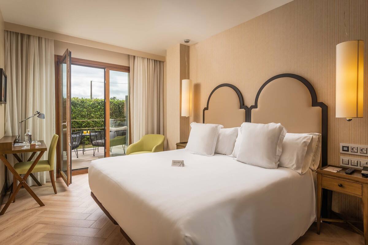 A hotel room with a large bed, a desk with a chair, a small table, and a balcony with a view of greenery.
