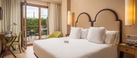 A hotel room with a large bed, a desk with a chair, a small table, and a balcony with a view of greenery.
