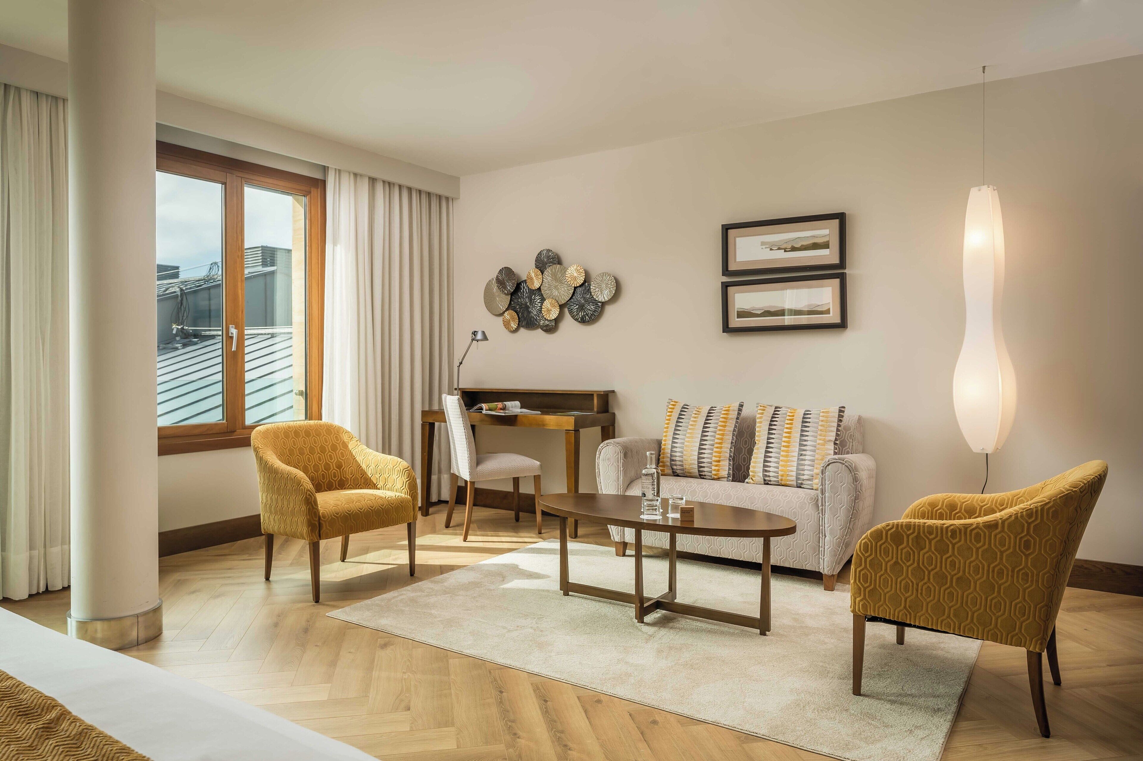 Un salon moderne comprenant un canapé, deux fauteuils, une table basse et un bureau. Il y a une grande fenêtre avec des rideaux, un luminaire fixé au mur et deux tableaux encadrés.