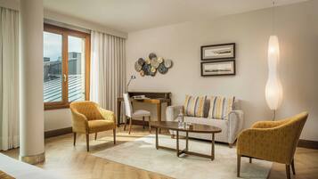 A modern living room with a sofa, two armchairs, a coffee table, and a desk. There is a large window with curtains, a wall-mounted light fixture, and two framed pictures.