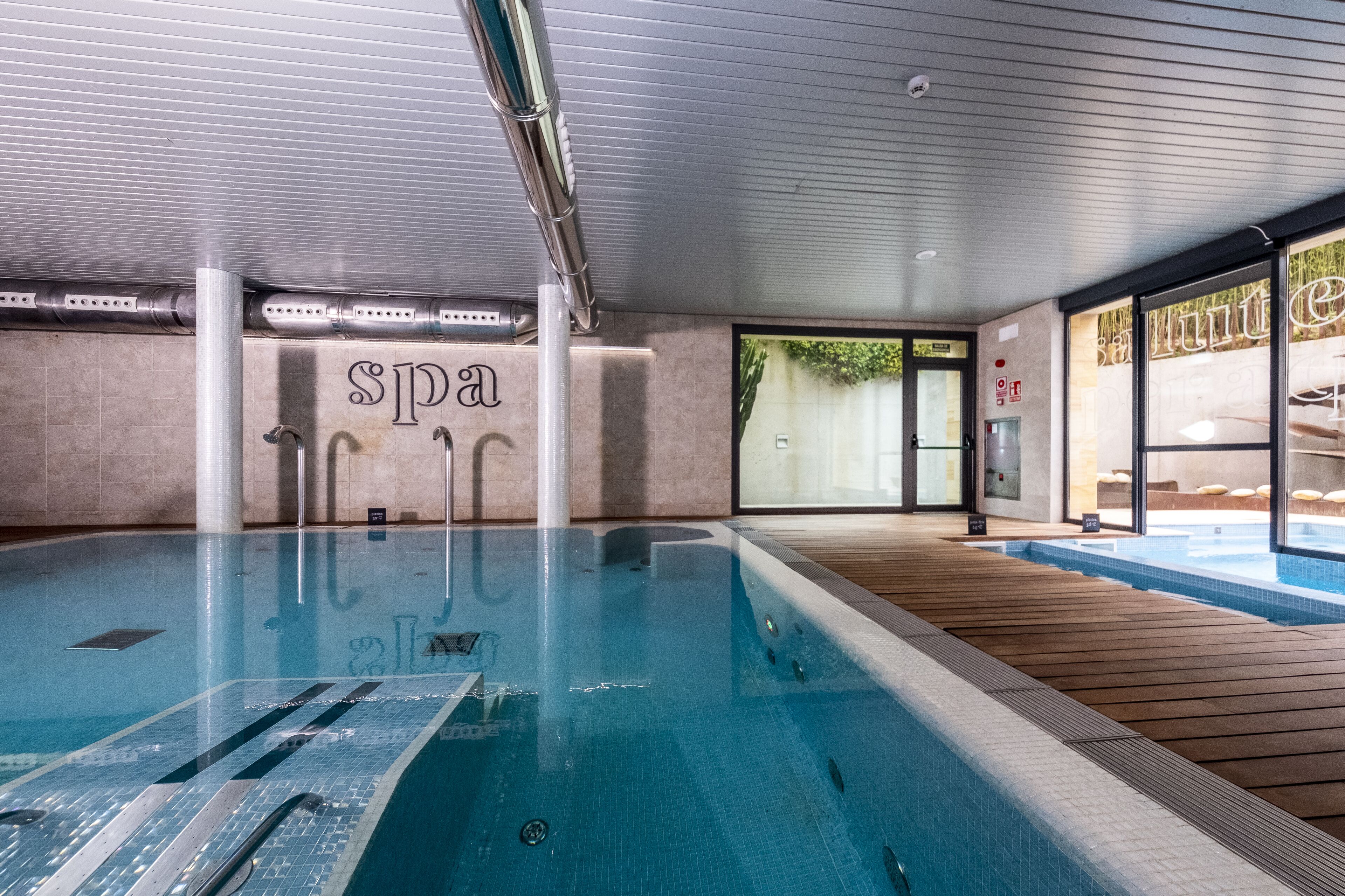 Una piscina interior con zona de spa, terraza de madera y vistas al exterior.