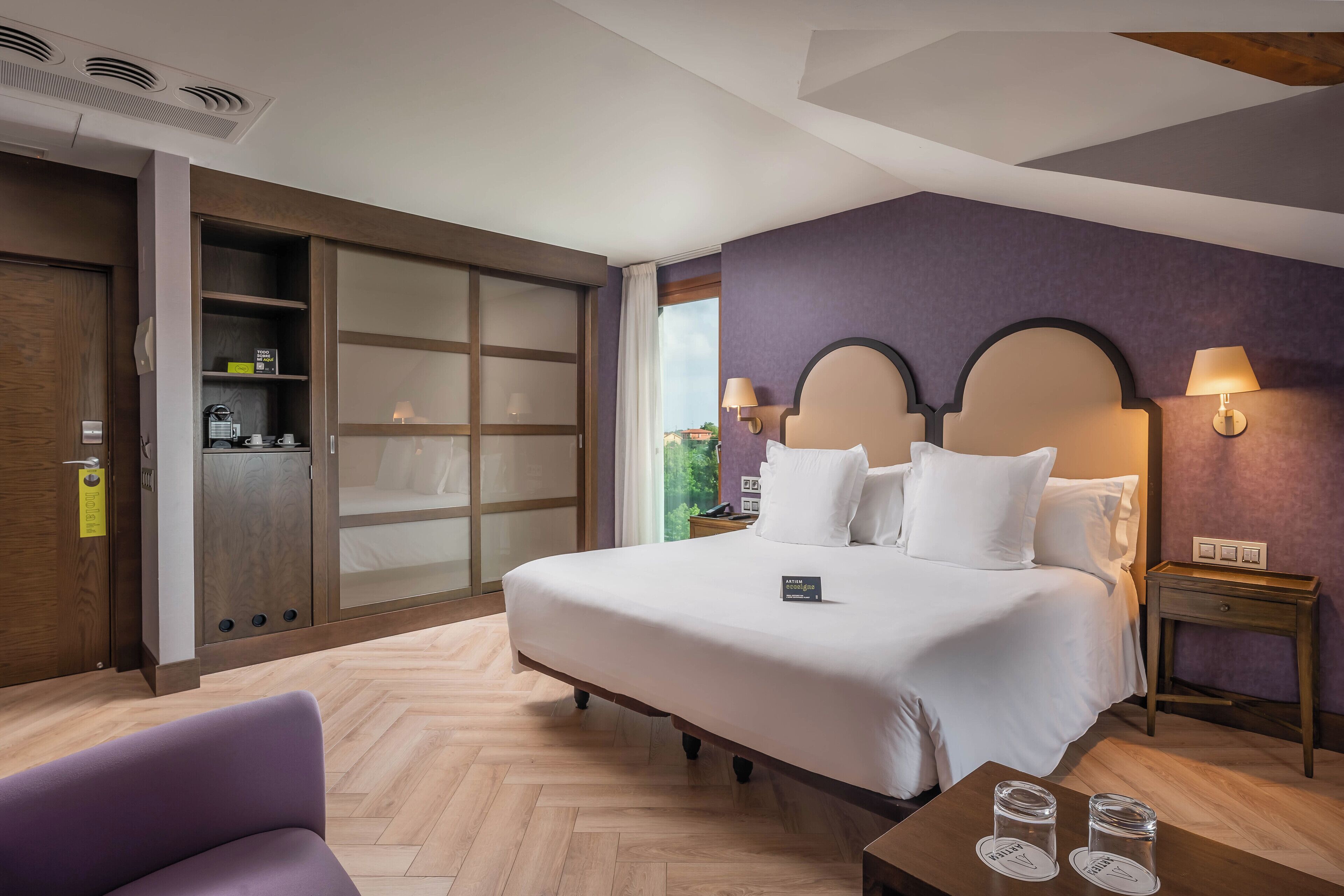 A hotel room with a large bed, a wardrobe with sliding doors, a bedside table with a lamp, and a view of greenery outside.