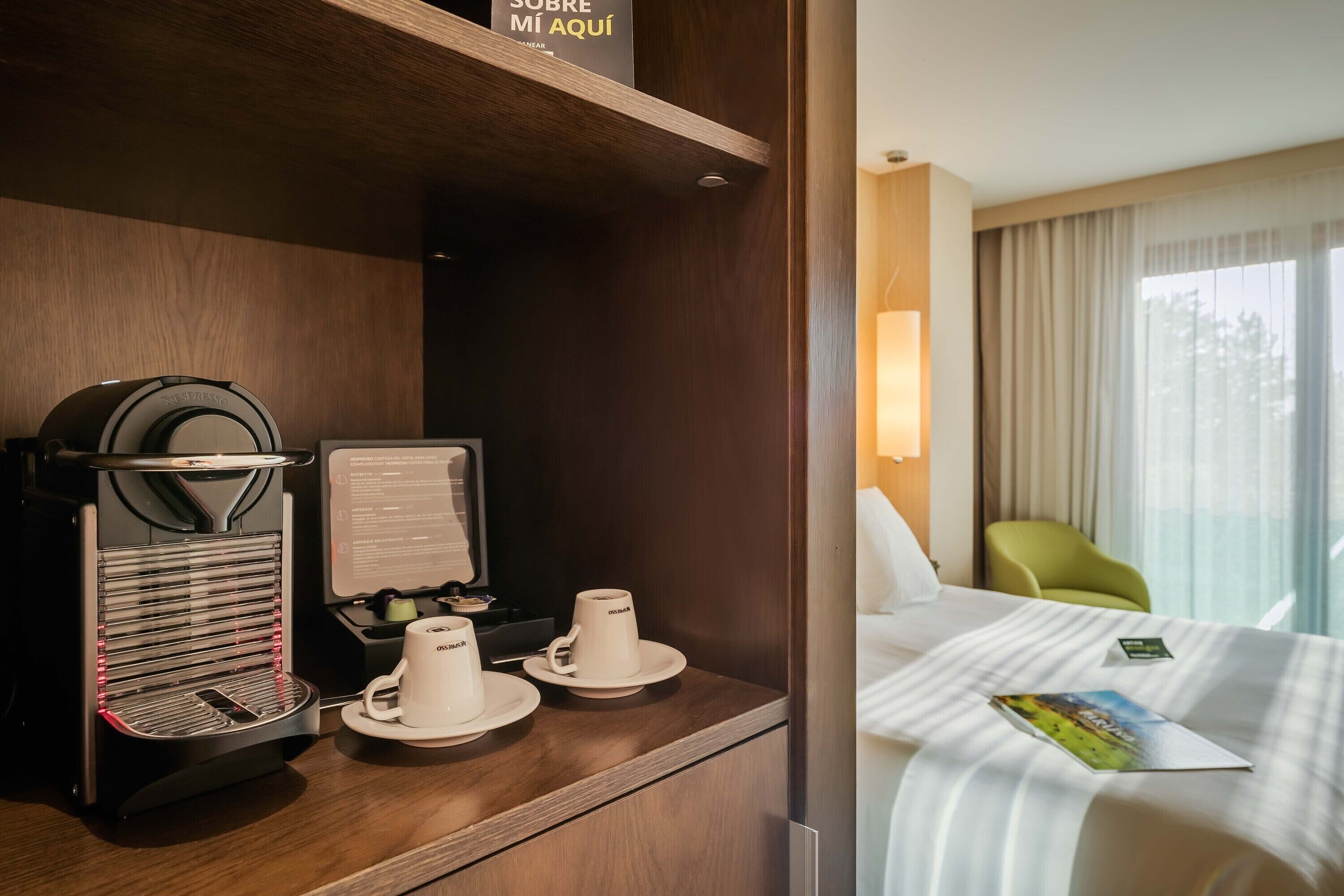 Une chambre d’hôtel avec un bureau en bois, une machine à café, deux tasses blanches, un lit recouvert d’une courtepointe à rayures, une chaise verte et une fenêtre avec des rideaux.