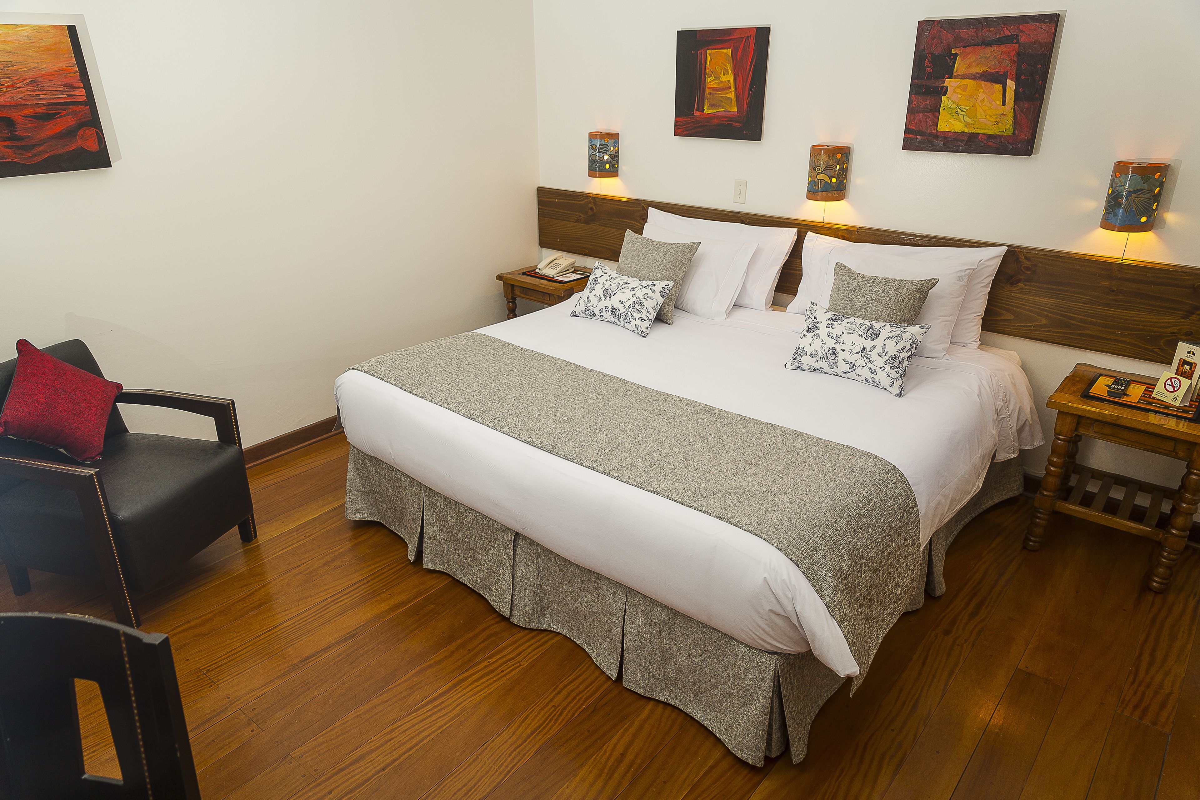 A neatly made bed with a wooden headboard, two bedside tables with lamps, a chair, and artwork on the walls.