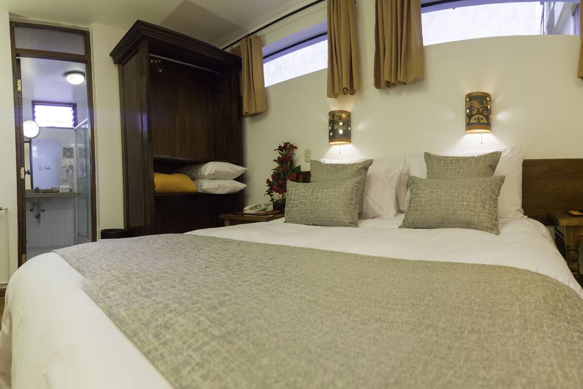 A hotel room with a large bed, a wooden headboard, a nightstand, a bedside table, a mirror, a wardrobe, and a window with curtains.