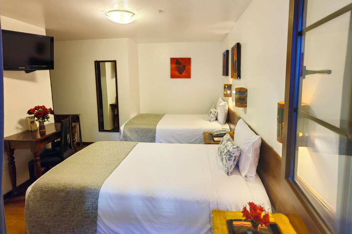 A hotel room with two beds, a desk with a chair, a flat-screen TV, a mirror, artwork on the wall, and a window with blinds.