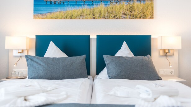 Ein Hotelzimmer mit zwei Betten, jeweils mit blauem Kopfteil, weißer Bettwäsche und Blick auf Strand und Steg durch ein großes Fenster.