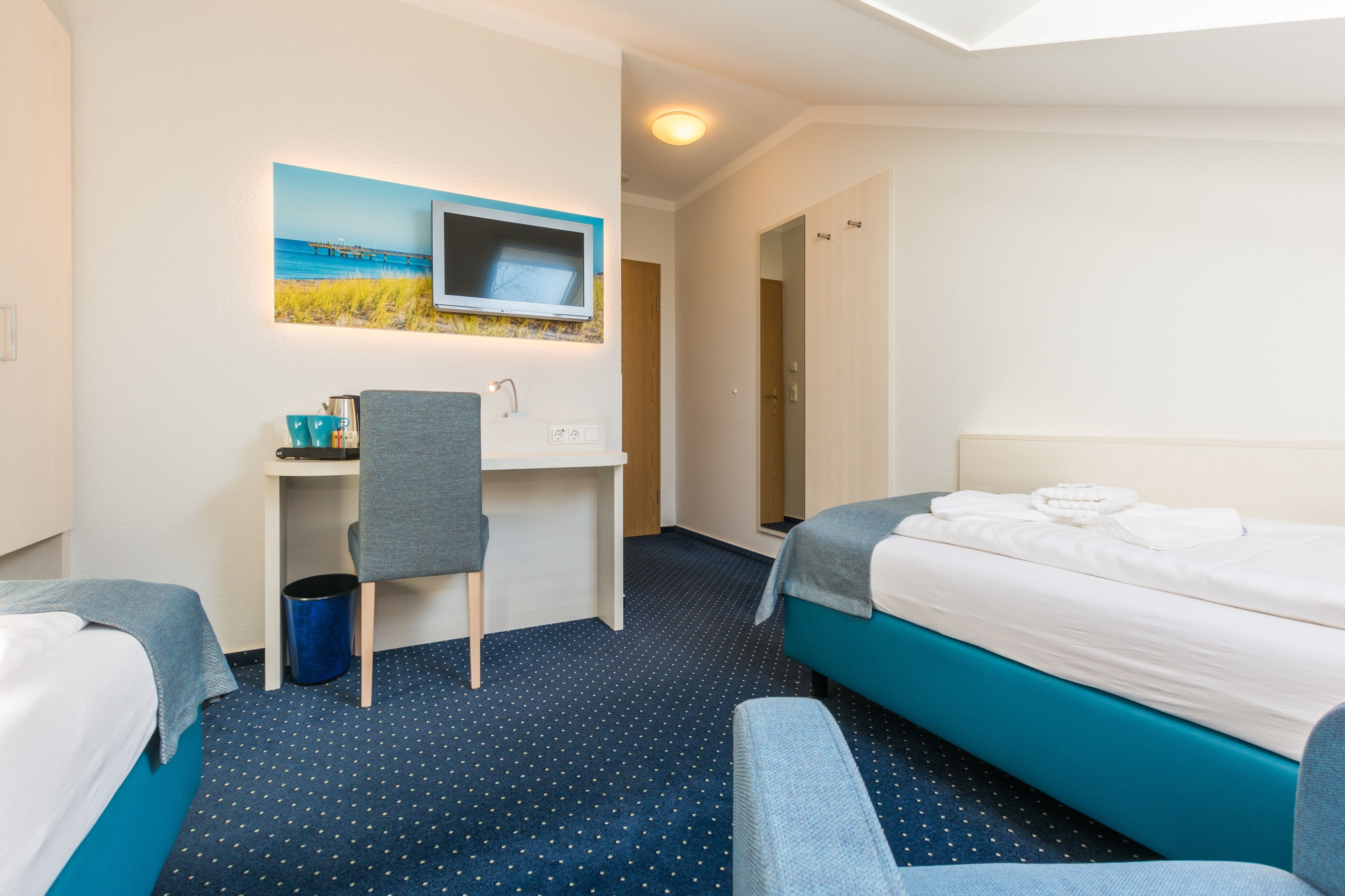 A hotel room with two beds, a desk with a chair, a TV mounted on the wall, and a painting of a beach scene.