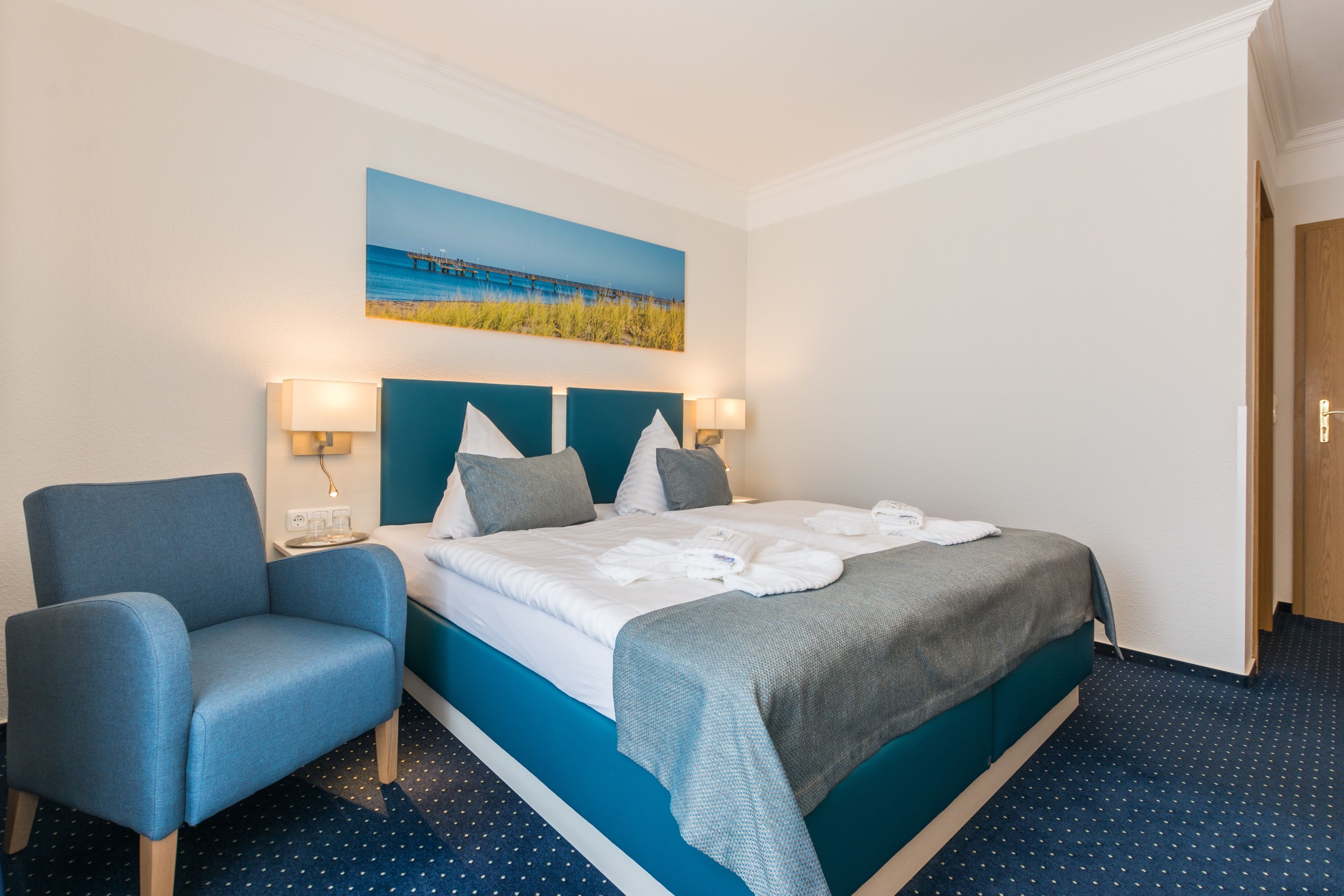 Ein Hotelzimmer mit einem Bett, zwei Nachttischlampen, einem Sessel und einem Bild von einer Strandansicht.