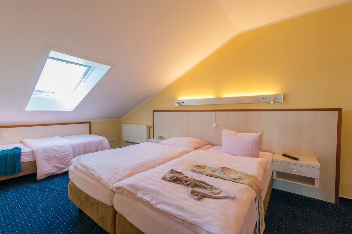 A hotel room with two beds, a skylight, a bedside table with a lamp, and a wall-mounted shelf.