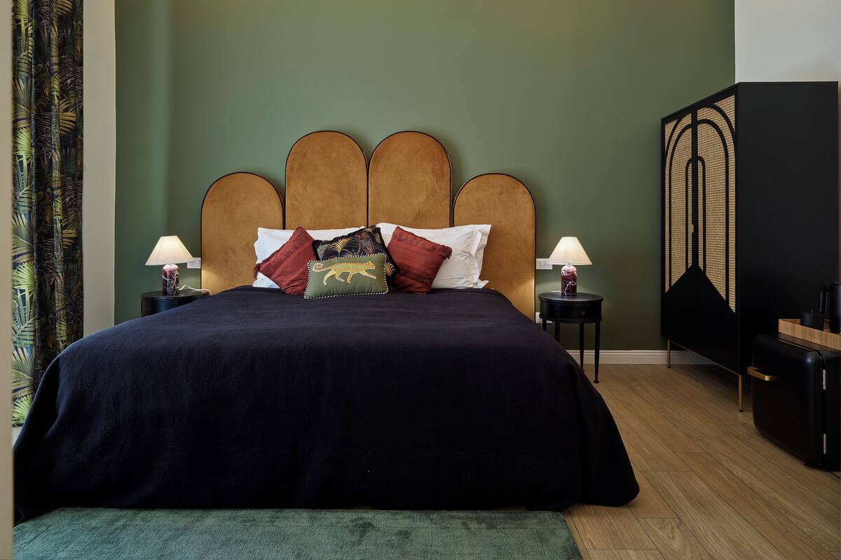 Een netjes opgemaakt bed met een donkerblauwe sprei en meerdere kussens, waaronder een opvallend kussen in de vorm van een dier. Twee nachtkastjes met lampen staan ernaast, en aan de rechterkant een grote, donkere kast.