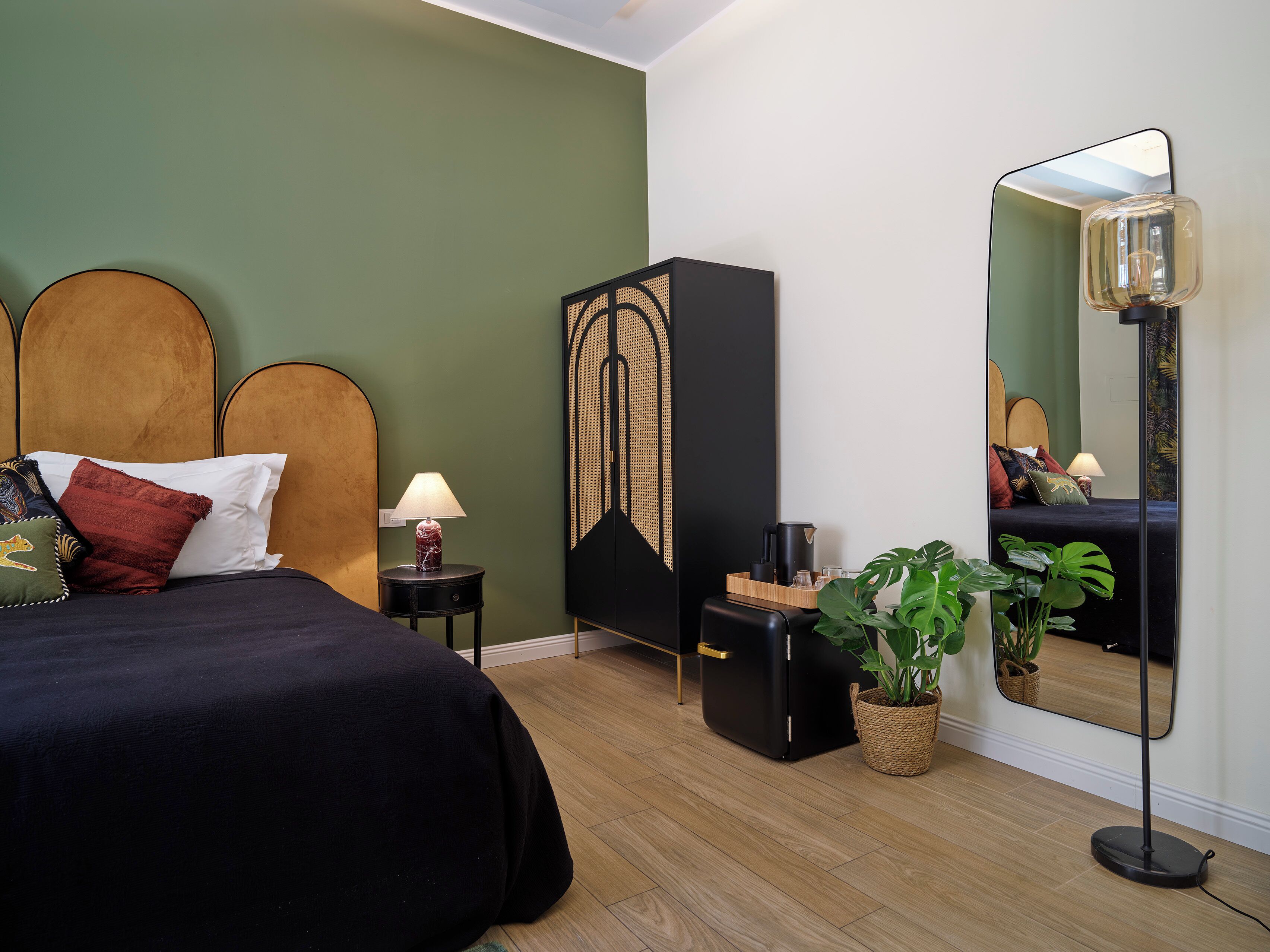 A bedroom with a bed, bedside table, lamp, wardrobe, mirror, and potted plants.