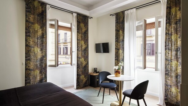 Une chambre d’hôtel avec un lit, une table ronde et deux chaises, un téléviseur fixé au mur et des rideaux à motifs de feuilles.