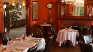 A dining area with tables set for two, wooden chairs, a large clock on the wall, and a fireplace.