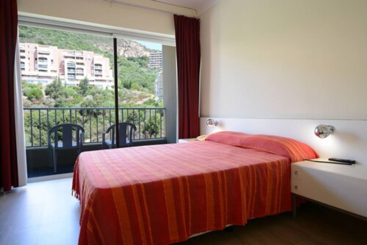 A bedroom with a bed, a nightstand, a window with a view of buildings and greenery, and a balcony with chairs.