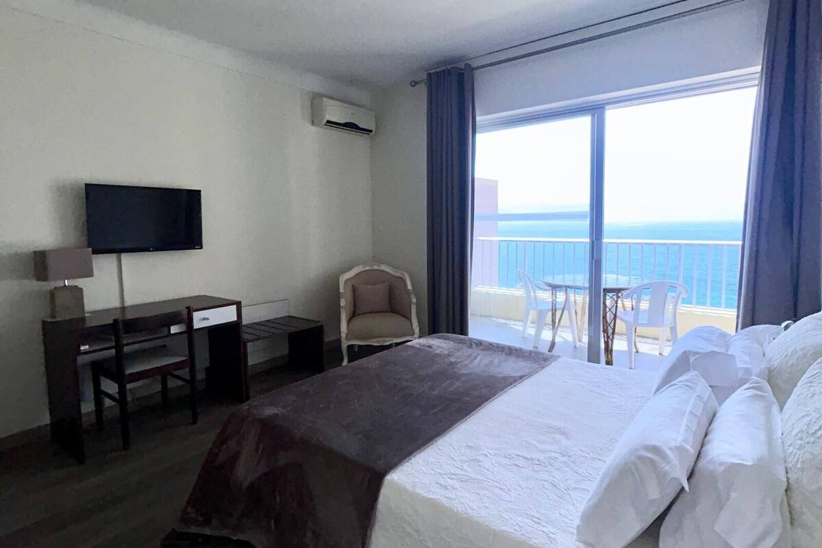 A hotel room with a bed, a desk, a chair, a television, and a balcony with a view of the sea.
