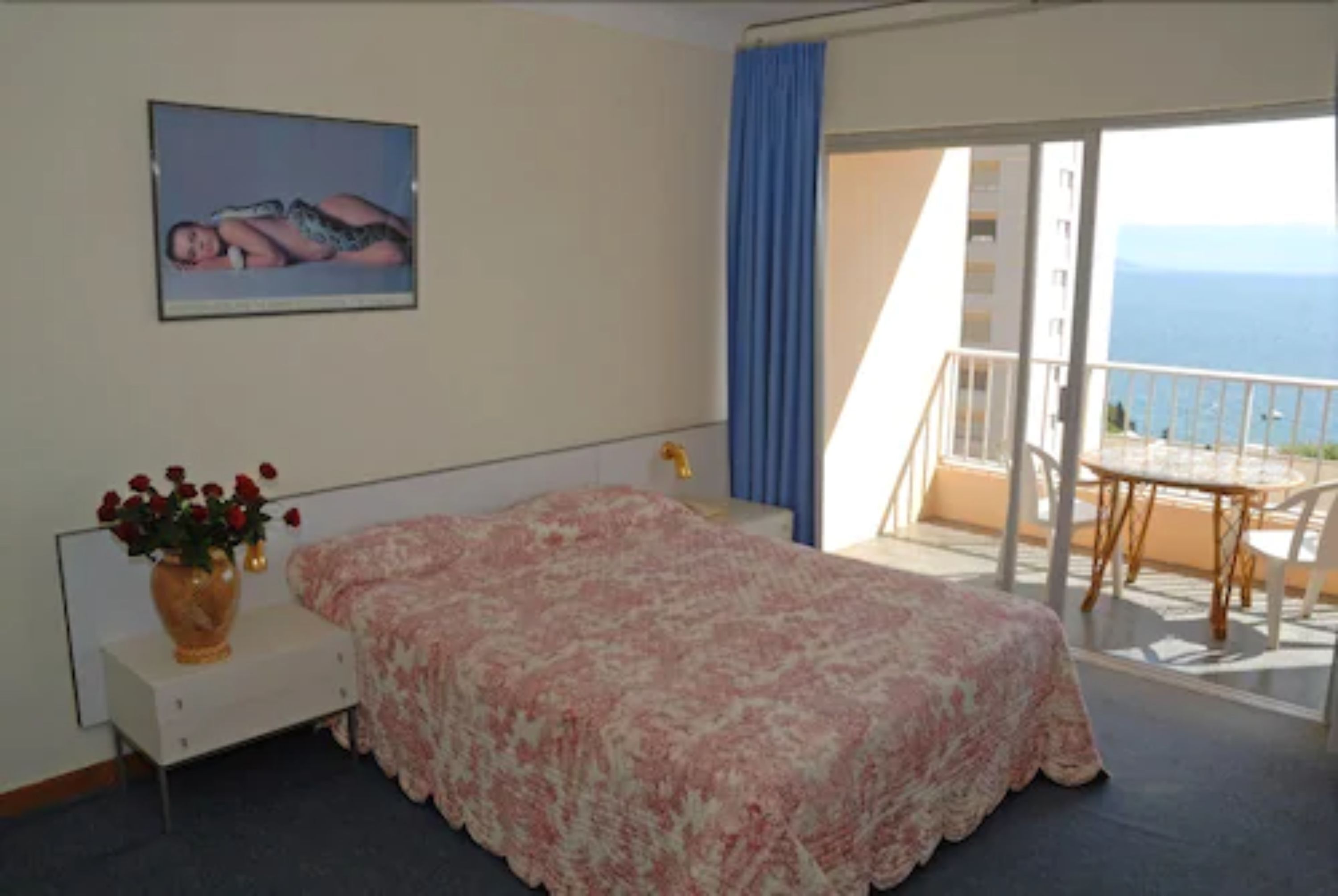 Une chambre à coucher avec un lit, une table de chevet ornée d’un vase de fleurs, une fenêtre donnant sur la mer et un balcon avec une table et des chaises.