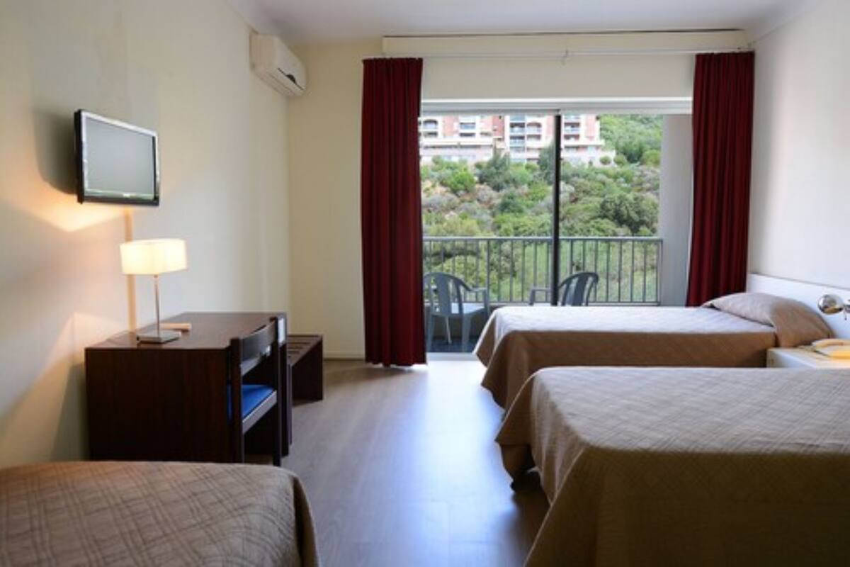 A hotel room with two beds, a desk with a lamp, a TV, and a balcony with a view of buildings and trees.